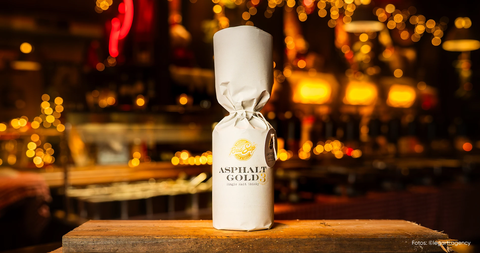 Verpackte Whiskyflasche „Asphalt Gold“ mit Packagingdesign auf Holzfläche vor Barhintergrund