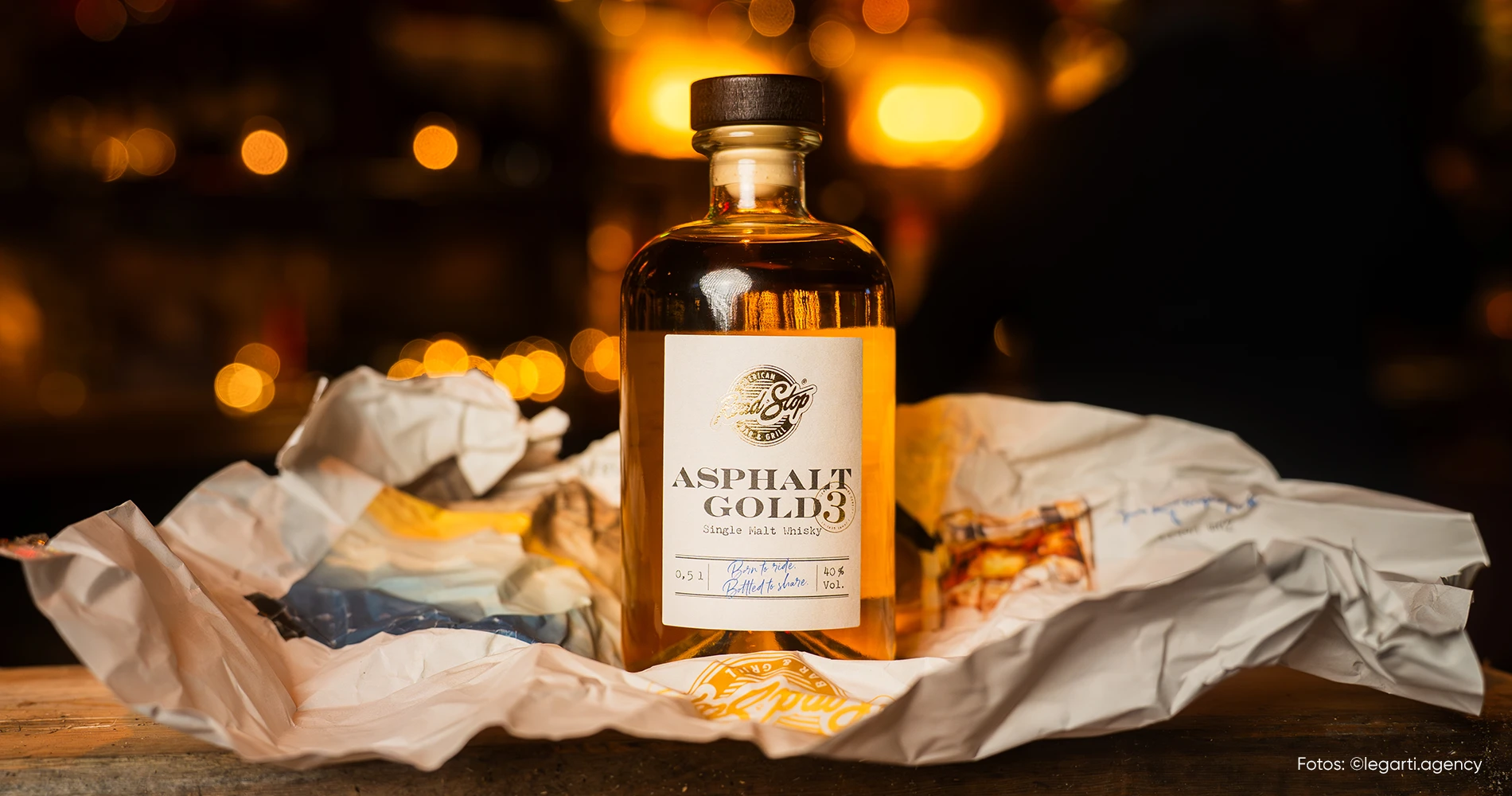 Whiskyflasche mit Packagingdesign „Asphalt Gold“ auf rustikalem Setting mit warmem Licht