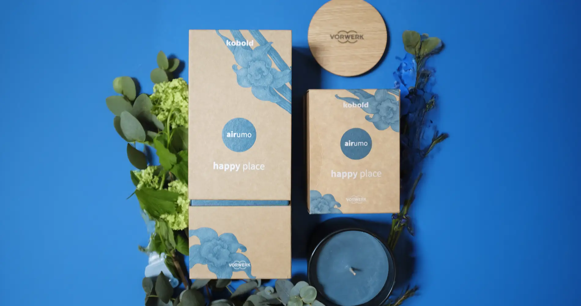 Produktinszenierung des Kobold Airumo Dufts „Happy Place“ mit Verpackung, Kerze und Pflanzen in blauer Farbwelt