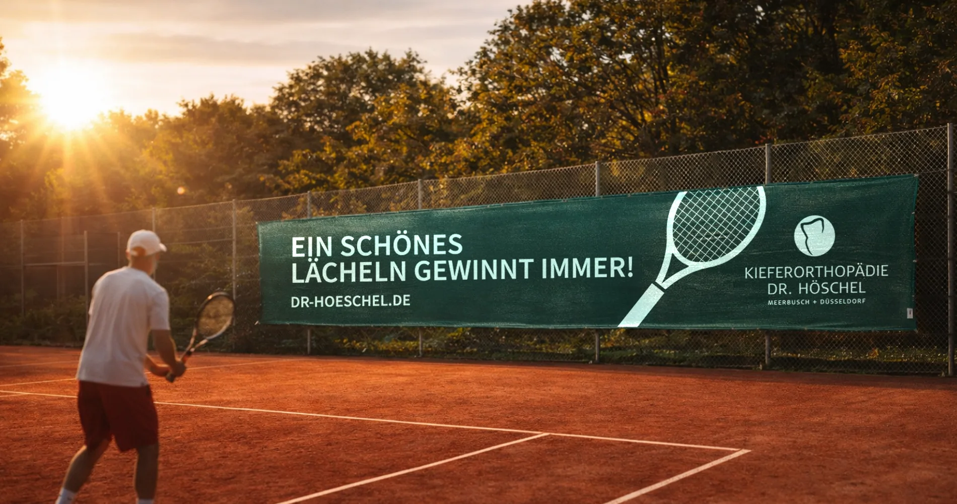 Tennisplatz mit Werbebanner für Dr. Höschel Kieferorthopädie, Spieler im Vordergrund, Claim „Ein schönes Lächeln gewinnt immer“