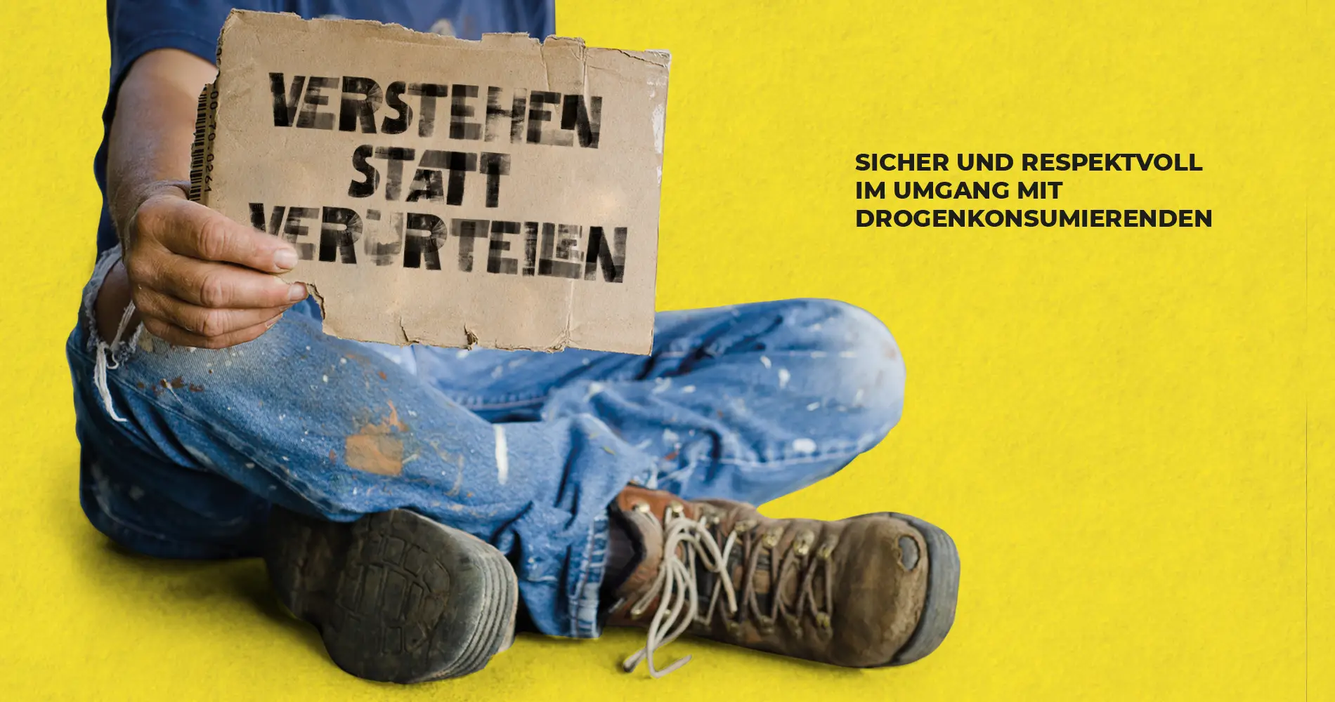 Person sitzt am Boden und hält Schild „Verstehen statt verurteilen“ als Kampagnenmotiv der Suchthilfe
