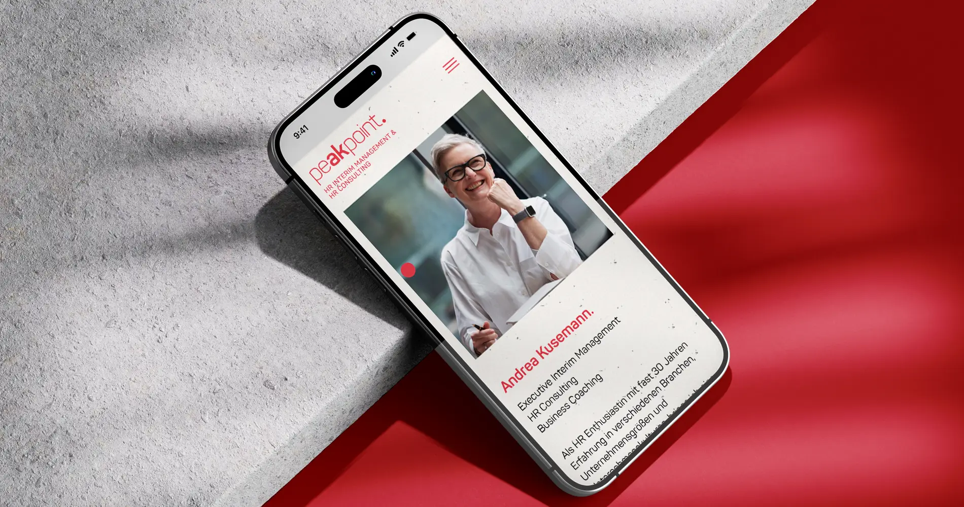 Smartphone mit Peakpoint Website auf dem Display, zeigt Profilseite mit Porträt und Text auf rot-grauem Hintergrund