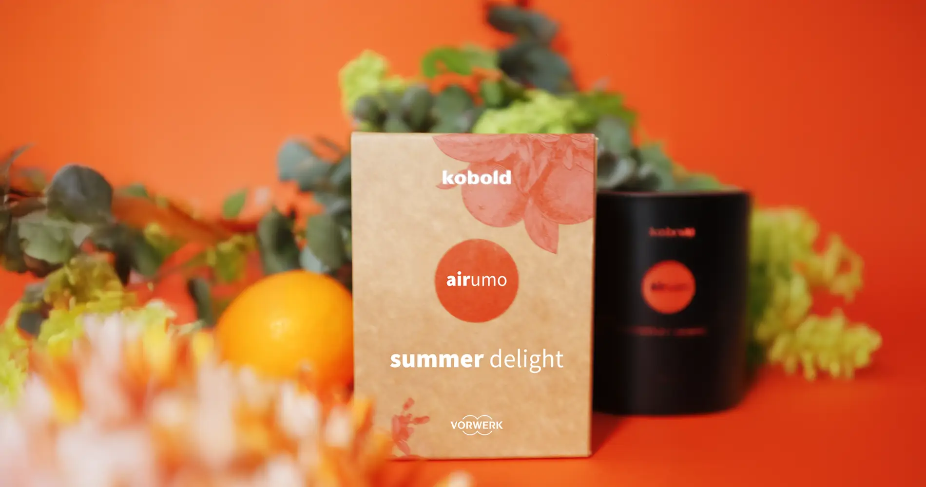 Produktinszenierung des Kobold Airumo Dufts „Summer Delight“ mit Verpackung, Diffusor und frischen Zitrus- und Blütenelementen vor orangem Hintergrund
