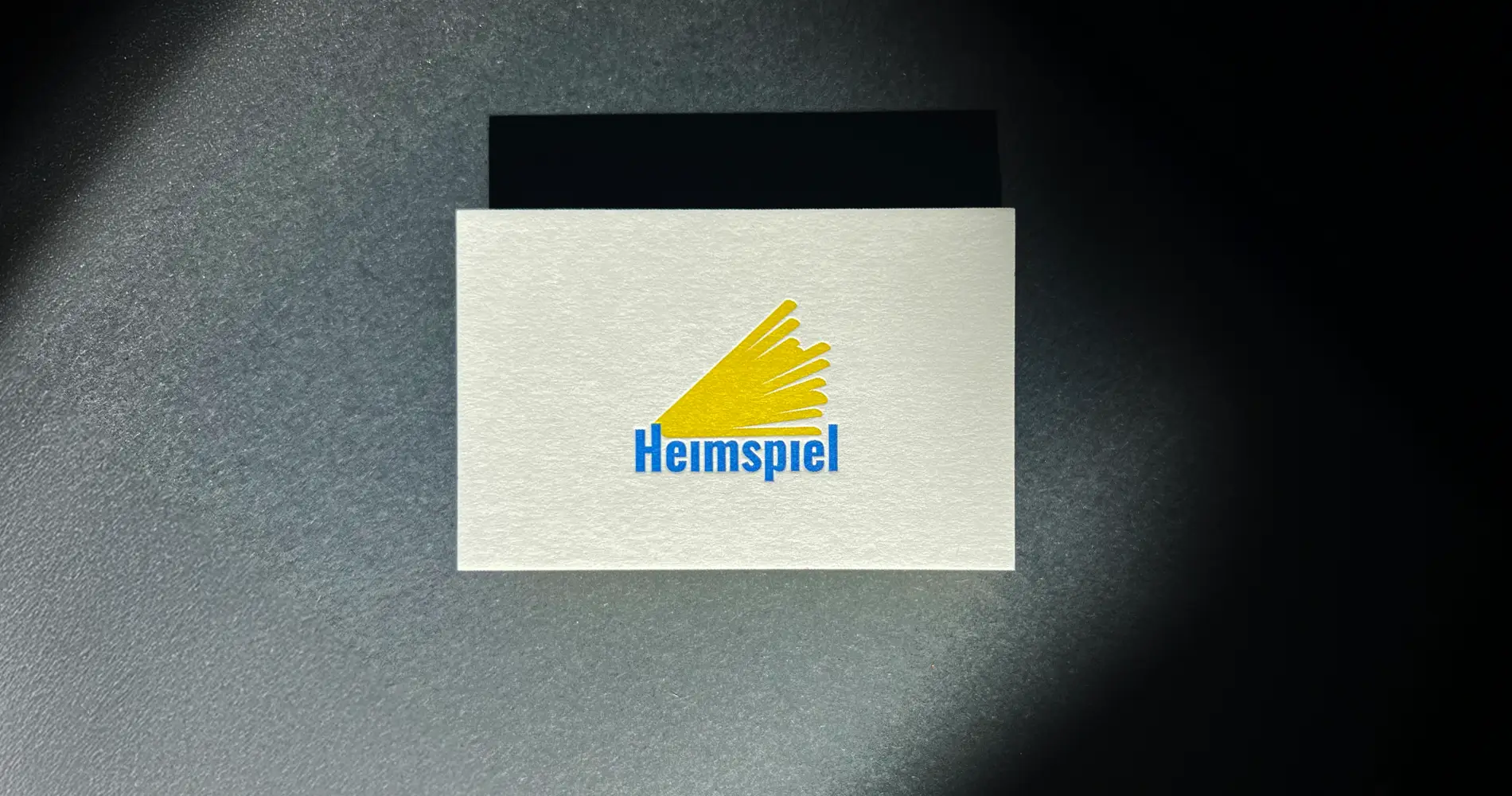 Visitenkarte mit Heimspiel-Logo im Licht auf dunklem Hintergrund